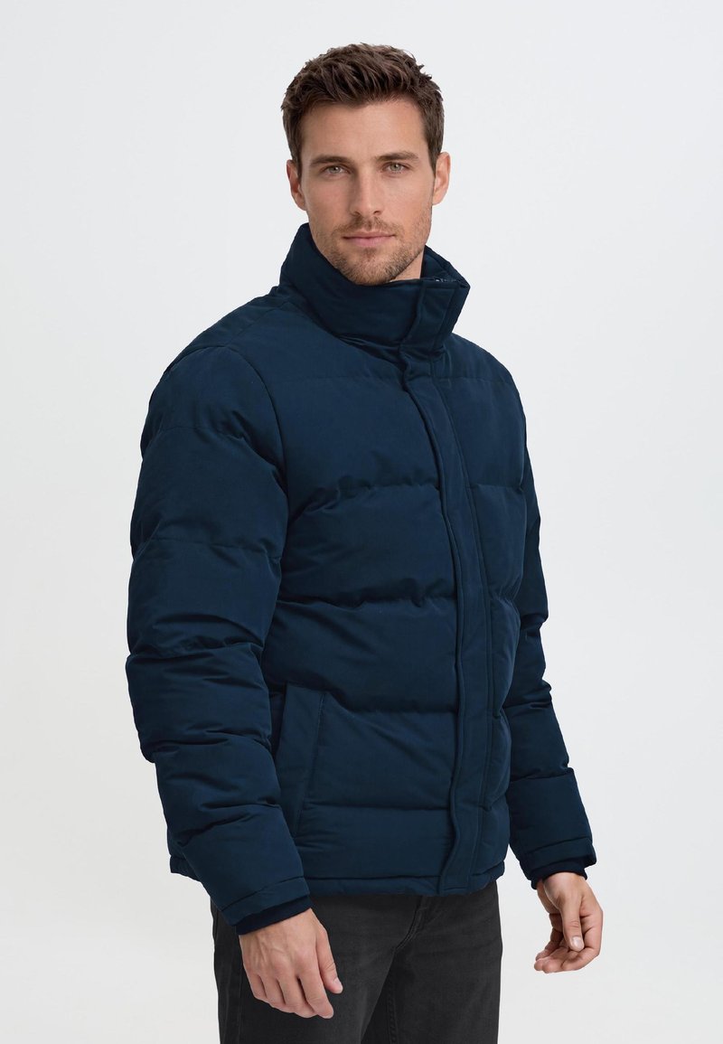 Mann mit kurzen braunen Haaren, der eine dunkelblaue Steppjacke trägt und vor einem schlichten hellen Hintergrund steht.