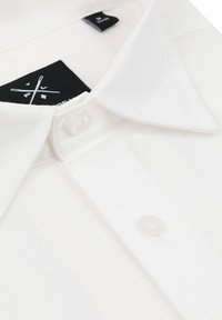 Biała bawełniana koszula polo z klasycznym kołnierzykiem, przezroczystymi guzikami i czarną metką wewnątrz. Gładka tekstura bez widocznych wzorów.