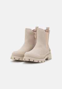 Botas de tobillo beige con parte superior de tela texturizada, laterales elásticos, detalle de cremallera dorada y suela robusta con tacos para mayor tracción.
