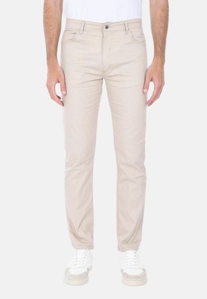 Uomo che indossa pantaloni beige chiaro aderenti e sneakers bianche, in piedi davanti a uno sfondo chiaro e semplice.