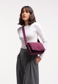 Borsa a spalla imbottita color mogano con chiusura a pattina e tracolla regolabile. Texture liscia, forma rettangolare, abbinata a un top bianco aderente e pantaloni grigi.