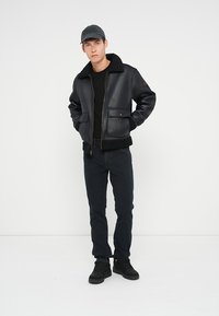 Svart lær bomberjakke med saueskinnskrage, glidelås foran og to sidelommer. Brukt med en svart t-skjorte og mørke jeans.