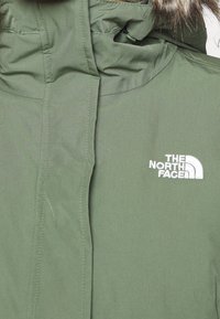 Veste d'hiver verte avec une capuche bordée de fourrure, avec le logo "The North Face" brodé en blanc sur la poitrine.