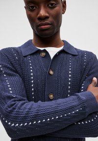 Sötétkék kötött kardigán texturált mintával, két barna gombbal, galléros nyakkal és nyitott szövésű mintákkal az ujjakon.