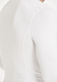 T-shirt de sport à manches longues blanc avec une finition lisse, présentant des détails en mesh sur le côté et des coutures plates pour le confort.