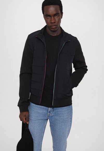 Svart hettejakke med quiltet front, fargede glidelåsdetaljer, lange ermer og tettsittende mansjetter, sammen med en svart skjorte og blå jeans.