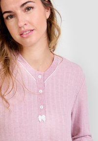 Mujer vistiendo una blusa de manga larga de color rosa con relieve, con botones blancos y un pequeño lazo blanco cerca del cuello, sobre un fondo claro y liso.