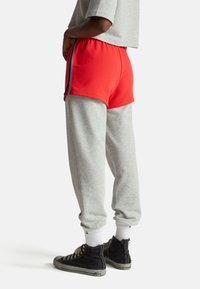 Osoba nosící červené šortky přes šedé tepláky, černé vysoké tenisky, bílé ponožky a šedý krátký top, stojící bokem na bílém pozadí.