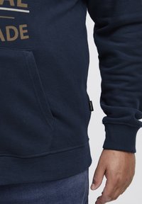 Felpa in cotone blu navy con tasca anteriore, polsini e orlo a costine. Presenta dettagli in testo dorato sul petto. Piccolo tag del marchio visibile.