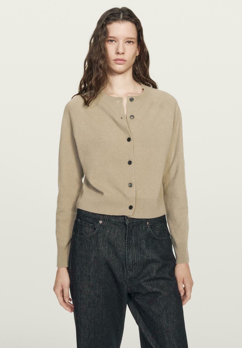 Jonge vrouw gekleed in een beige gebreid vest met knopen en een donkere, hooggetailleerde spijkerbroek, staand tegen een effen lichte achtergrond.