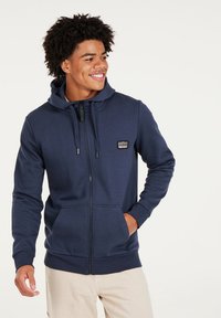 Navy zip-up hoodie in soft fabric with adjustable drawstring hood, two front pockets, and a small logo patch on the chest.