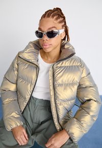 Giacca puffer metallica dorata con colletto alto e trapuntatura prominente; abbinata a un top bianco a coste e pantaloni verdi, con occhiali da sole ovali.