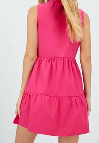 Vaaleanpunainen hihaton mekko, jossa on kerroksellinen röyhelöhima ja kaulus, joka tarjoaa sileän tekstuurin ja ryhdikkään muotoilun. Kirkkaat ompeluyksityiskohdat.