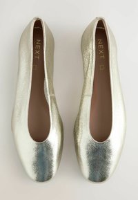 Bailarinas metálicas plateadas con una textura suave, punta redondeada y lados recortados. Nombre de la marca "NEXT" impreso en el interior.