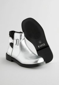 Botas de tobillo metálicas plateadas con un acento de lazo, lados elásticos negros y suela de goma. Textura suave y diseño de punta redonda.