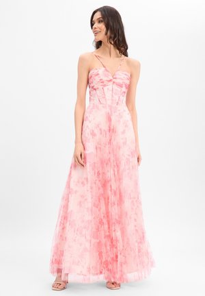 Vrouw in een vloerlange roze bloemenjurk met spaghetti bandjes, glimlachend en opzij kijkend tegen een effen witte achtergrond.