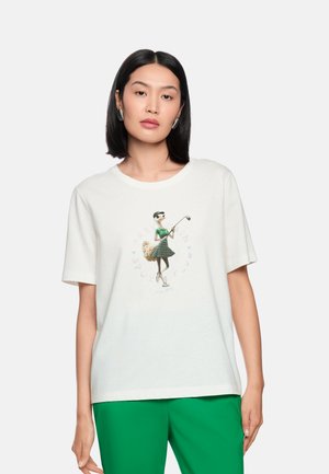 Woman with black hair wearing white T-shirt with graphic of an animal playing golf and green pants, standing against a plain background.