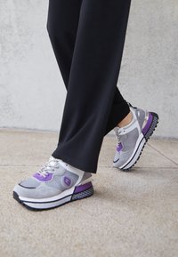 Le sneaker grigie presentano materiali in rete e suede con accenti viola, hardware argento e suole nere testurizzate per una migliore aderenza.