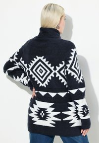 Maglione lavorato a maglia con colletto alto, caratterizzato da un motivo geometrico blu navy e bianco, maniche lunghe e una vestibilità morbida. Tessuto testurizzato con cuciture visibili.