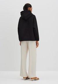 Felpa nera con cappuccio e vestibilità comoda, abbinata a pantaloni larghi beige chiaro e scarpe aperte con tacco basso. Visualizzazione da dietro.