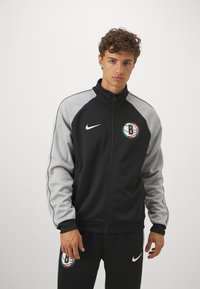 Μαύρο μπουφάν με φερμουάρ και γκρι μανίκια, με λογότυπο μπάσκετ στο αριστερό στήθος και λευκή λωρίδα Nike στο μπροστινό μέρος. Απαλή υφή υφάσματος.