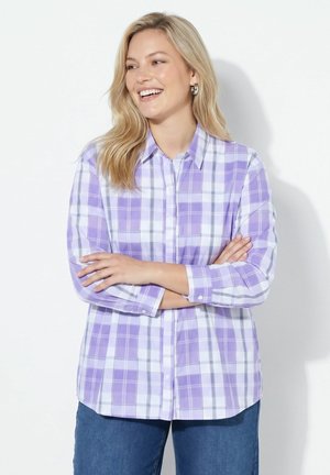 Donna sorridente con lunghi capelli biondi che indossa orecchini a cerchio d’argento, una camicia a quadri viola e bianca e jeans blu con le braccia incrociate.