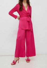 Vrouw in helderroze satijnen wijde broek met bijpassende gebelte blazer en hoge sandalen versierd met roze veren.