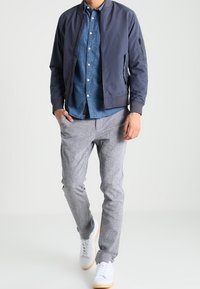 Man walking wearing a navy bomber jacket, blue button-up shirt, gray pants, and white sneakers against a plain background.