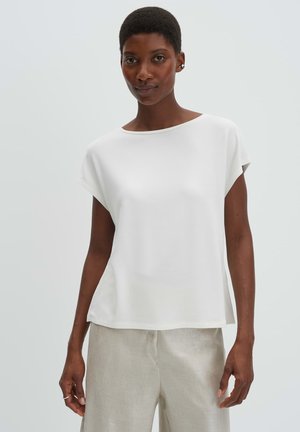 Woman with short hair wearing a white short-sleeve top and beige pants, standing against a plain light background.