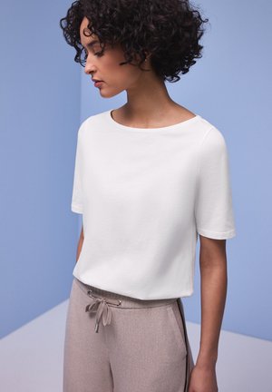 Kobieta o kręconych ciemnych włosach nosi gładką białą koszulkę z krótkim rękawem i jasnobrązowe spodnie ze ściągaczem na tle niebieskiego tła.