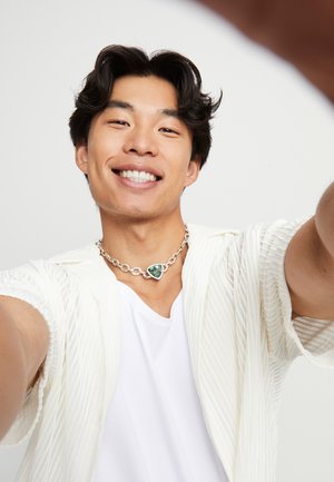 Collier chaîne argentée avec un pendentif triangulaire vert, porté sur un t-shirt blanc. La personne porte une chemise blanche ouverte à texture.