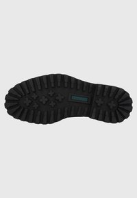 Suela de zapato de goma negra con un patrón de relieve profundo, que presenta tachuelas circulares y rectangulares para tracción, y un pequeño logo en el centro.