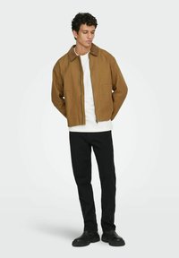 Mladenič stoji s rokami za hrbtom, oblečen v rjav zadrgo jakno, belo srajco, črne hlače in črne škornje ob ozadju brez vzorca.