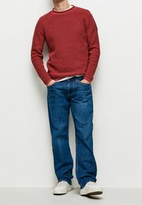 Roter strukturierter Strickpullover mit Rundhals, kombiniert mit blauen Jeans und weißen Turnschuhen. Die Designs sind lässig und in entspannter Passform.