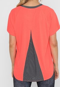 Krátký rukáv oranžový top s rozparkem na zadní straně, který odhaluje černobíle vzorovanou podšívku. Hladká textura s zaobleným lemováním.