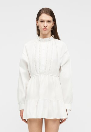 Vrouw draagt een witte mini-jurk met lange mouwen, kantdetails, een geplooide kraag en een geraffineerde taille, staand tegen een effen achtergrond.