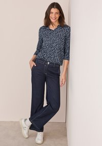 Blusa a fantasia navy con maniche a ¾, abbinata a jeans larghi in denim scuro e sneakers bianche. Tessuto morbido con vestibilità rilassata.