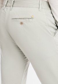 Helle graue Hose aus glattem Stoff, mit einer Gesäßtasche, die einen Holzknopf und dezente Nähte aufweist. Lässiger Schnitt.