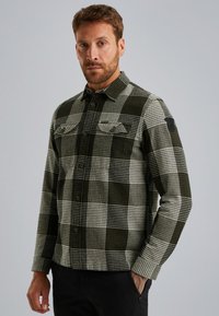 Chemise à carreaux dans les tons de vert foncé et crème, en tissu doux. Présente un devant à boutons, deux poches poitrine et un patch logo noir sur la manche.