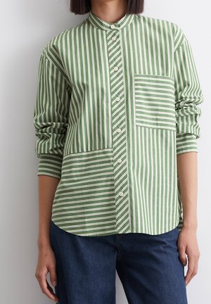 Frau trägt ein langärmeliges, grün-weiß gestreiftes Hemd mit Knöpfen und Brusttasche, kombiniert mit dunkelblauen Jeans.