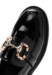 Mocassin en cuir verni noir avec détail mors clouté doré sur le dessus et bout arrondi sur fond blanc.