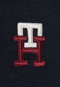 Geborduurd logo met verweven "H" in kastanjebruin en wit op een marineblauwe achtergrond. Textuur van gebreide stof.