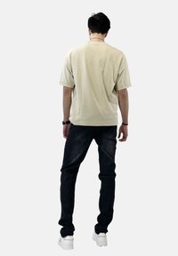 Camiseta beige de manga corta, de corte relajado, combinada con jeans de mezclilla oscuros. Modelo que lleva zapatillas blancas, visto desde atrás contra un fondo claro.
