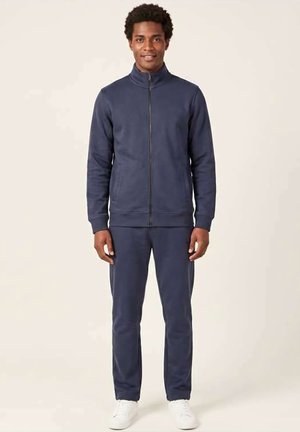 Mand står med fronten mod kameraet iført en navyblå lynlåstrøje, matchende bukser og hvide sneakers på en ensfarvet beige baggrund.