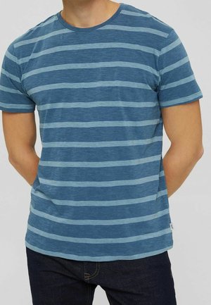 Mężczyzna ubrany w niebieski T-shirt z krótkim rękawem w jasnoniebieskie poziome paski oraz ciemne dżinsy, stoi z rękami za plecami.