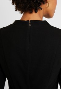 Frau mit kurzen Locken, die schwarze Kleidung mit einem sichtbaren Rückenzipper und einem kleinen goldenen Creolen-Ohrschmuck trägt.