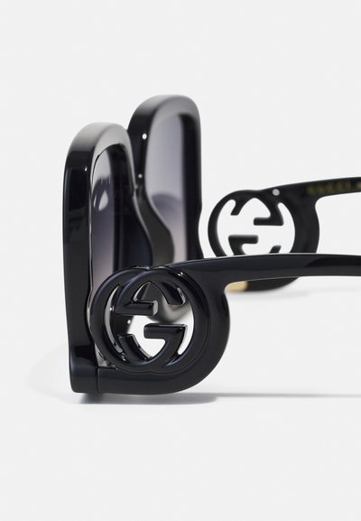 Gafas de sol negras con montura sobredimensionada, que presentan un logo circular prominente en las patillas, lentes degradados, acabado brillante y un diseño audaz.