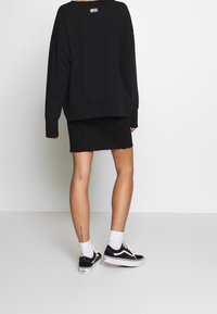 Sudadera negra de gran tamaño con puños acanalados, combinada con una falda vaquera negra con bordes deshilachados. Se lleva con zapatillas negras y blancas.