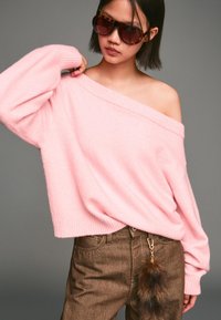 Rosa Off-Shoulder-Pullover aus weichem Strick, kombiniert mit braunen Hosen, die einen Fell-Pompom-Anhänger haben. Abgerundet mit großen, dunklen Sonnenbrillen.