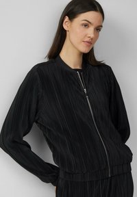 Veste bombardier noire avec plis verticaux, fermeture éclair sur le devant, col et poignets côtelés, et taille élastique, dotée d'un tissu lisse et texturé.